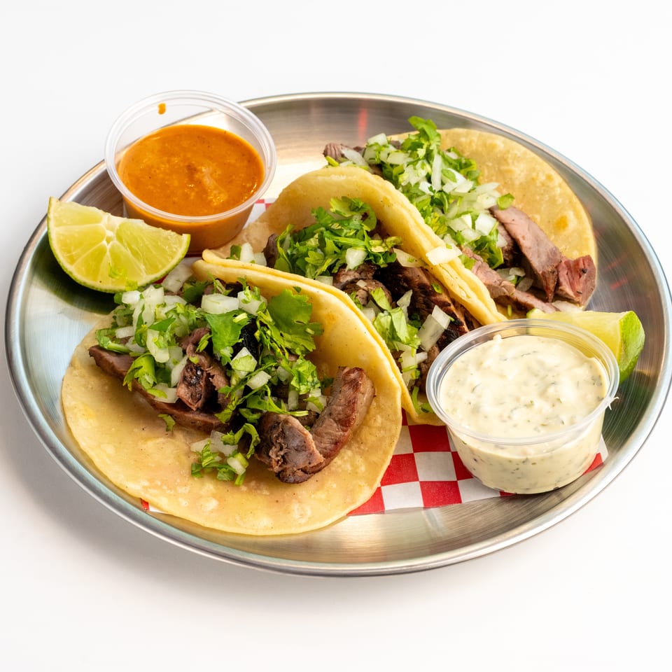 Three steak tacos with cilantro and onions, lime, and two sauces on a metal plate.