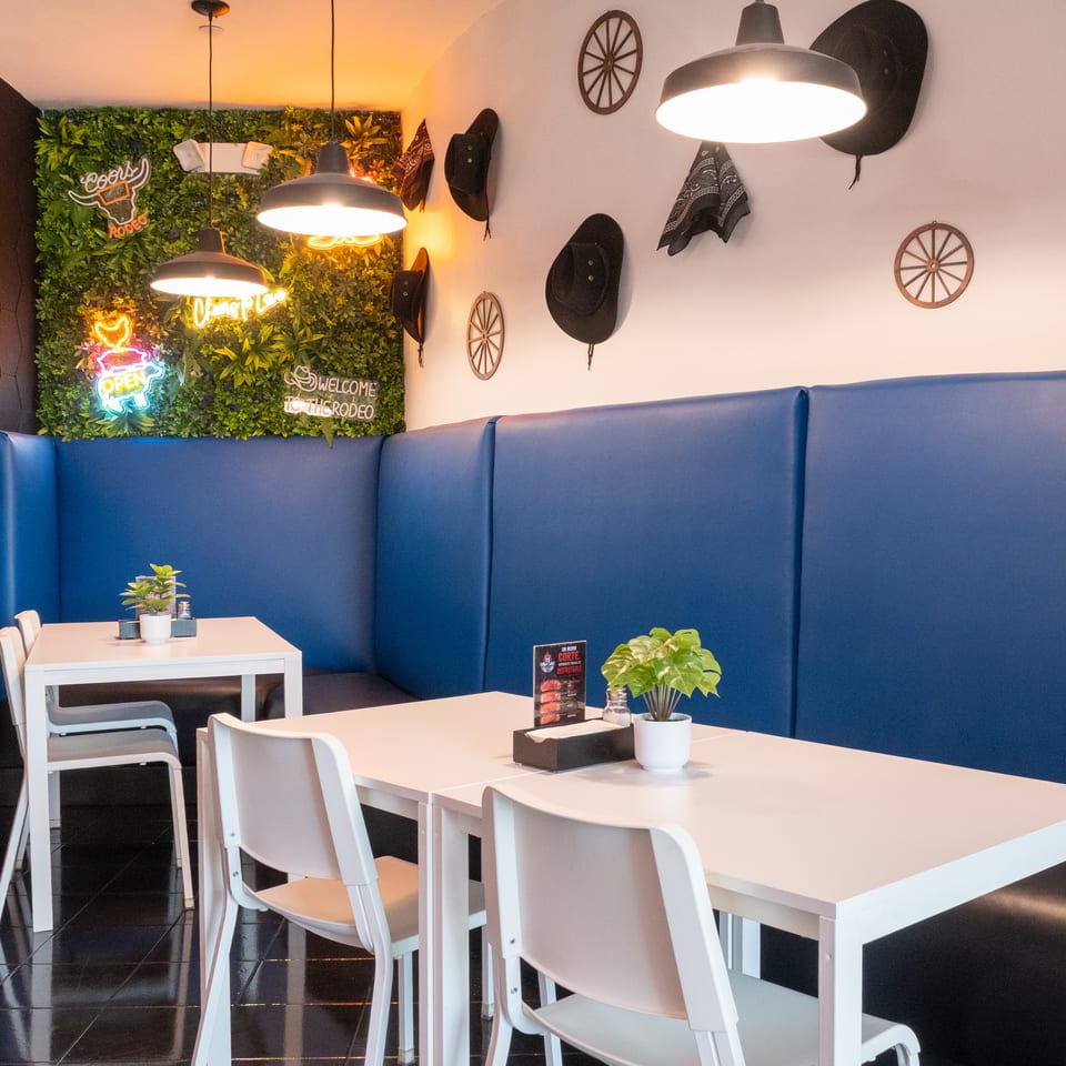 Restaurant interior with blue booth seating, white tables, and a green wall with neon signs and cowboy hats.