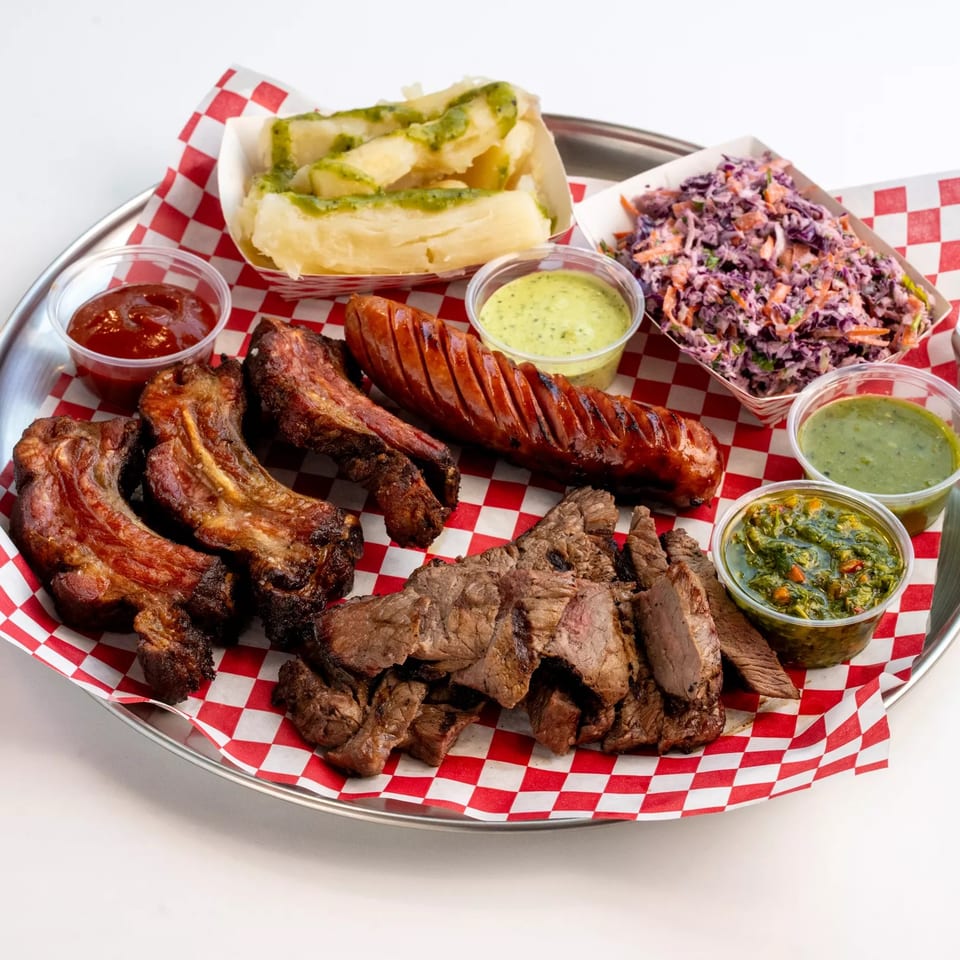Grilled meat platter with ribs, sliced steak, sausage, cassava, purple cabbage slaw, and assorted dipping sauces on checkered paper.