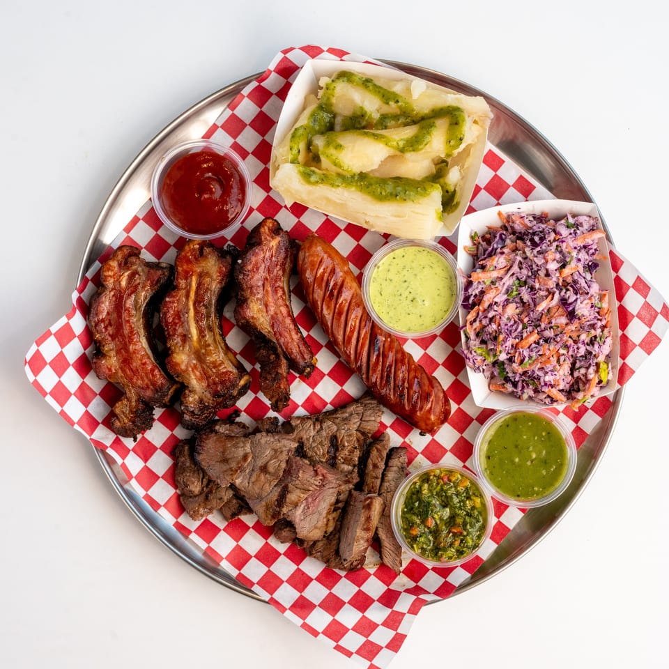 A platter of grilled ribs, sausage, steak, yucca, coleslaw, and various dipping sauces.