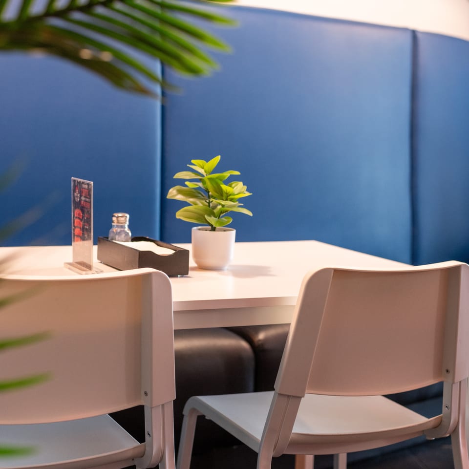 A restaurant table with two chairs, a plant, and condiments against a blue booth.