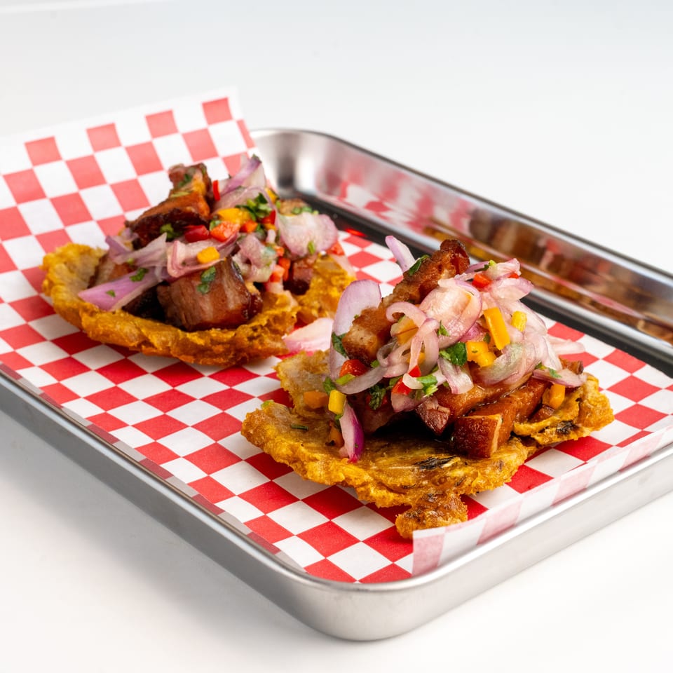 Two crispy plantain cups filled with savory pork belly, red onions, and colorful peppers on a tray.