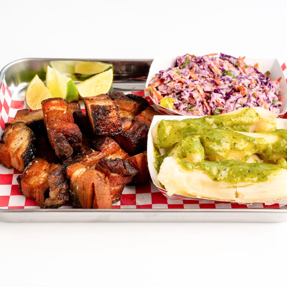Crispy pork belly chunks with lime wedges, coleslaw, and yuca with green sauce on a tray.