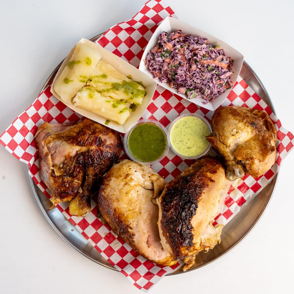 A platter of roasted chicken quarters, yucca, coleslaw, and two green sauces on a red checkered liner.