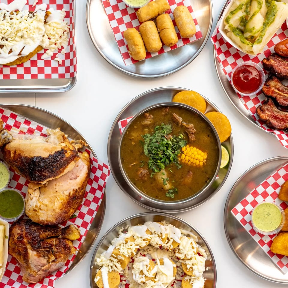 Overhead spread of Latin American dishes, including grilled chicken, ribs, fried pastries, soup, plantains, and sauces on checkered trays.