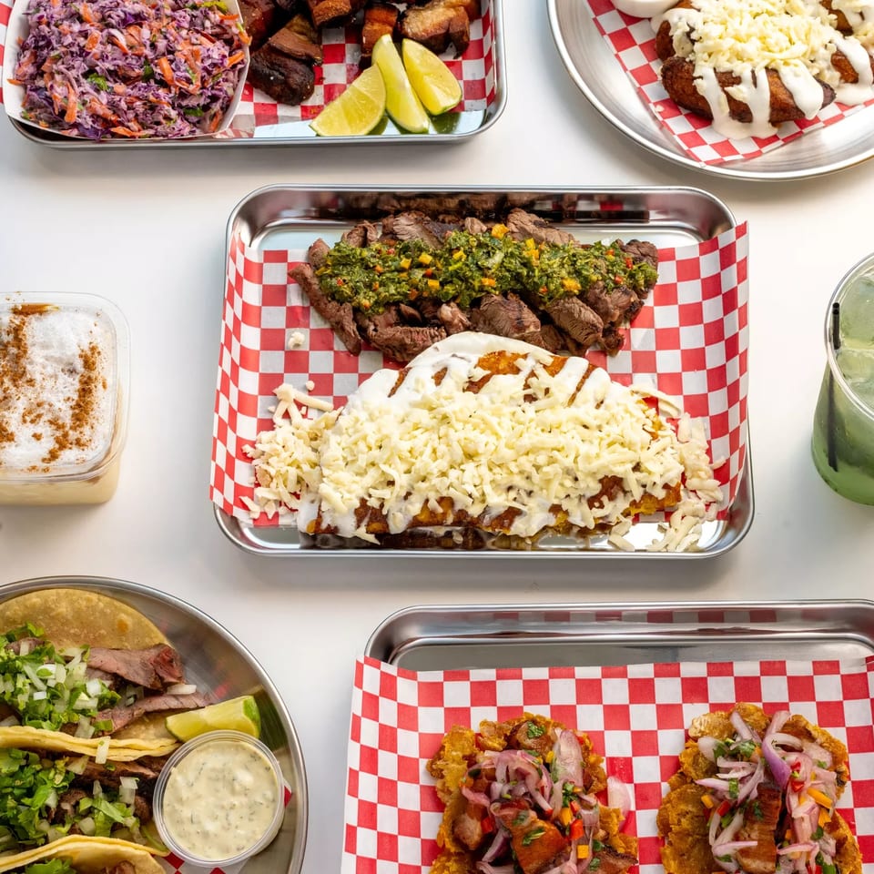 Overhead spread of Latin American dishes, including grilled steak, cheesy fries, tacos, slaw, lime wedges, and refreshing drinks.