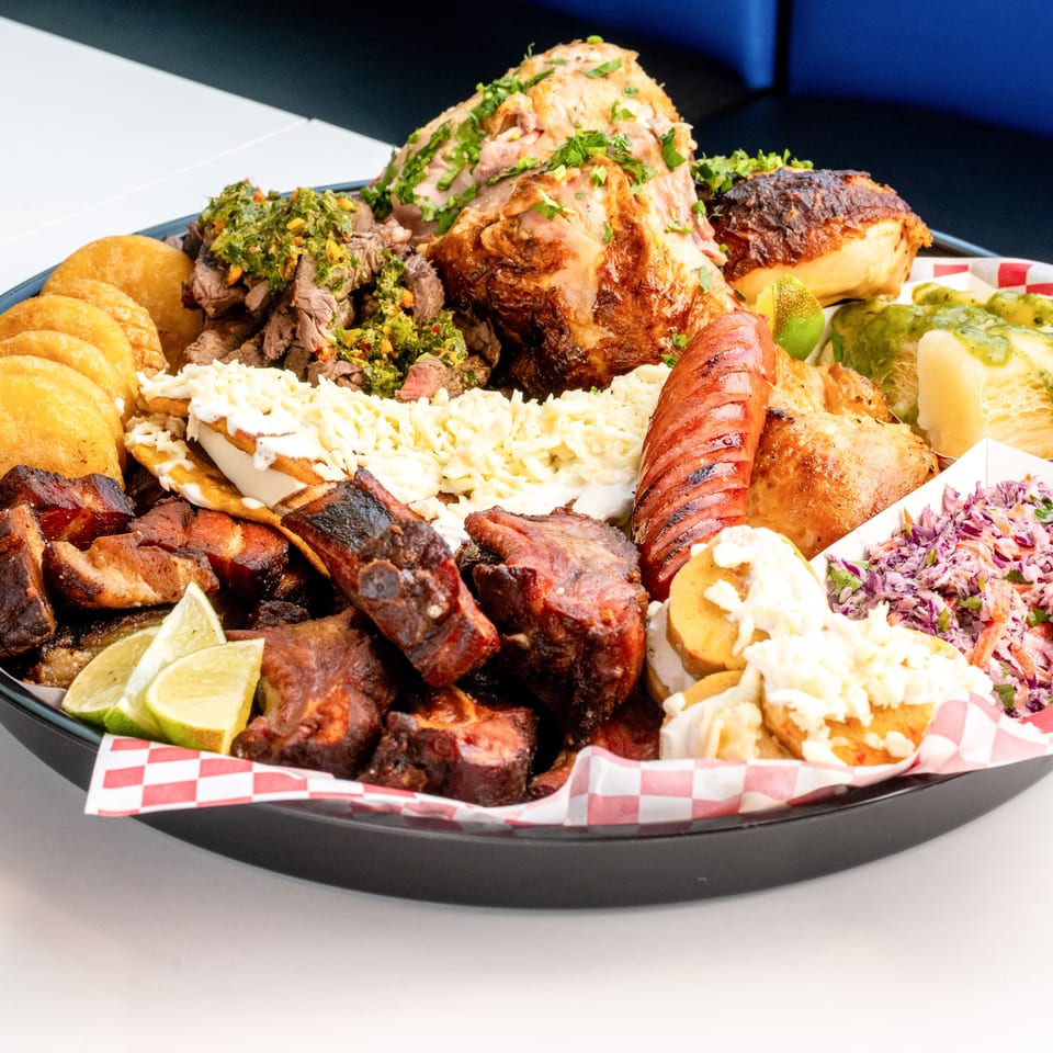 A large platter of various meats, empanadas, coleslaw, and other sides on a white table.