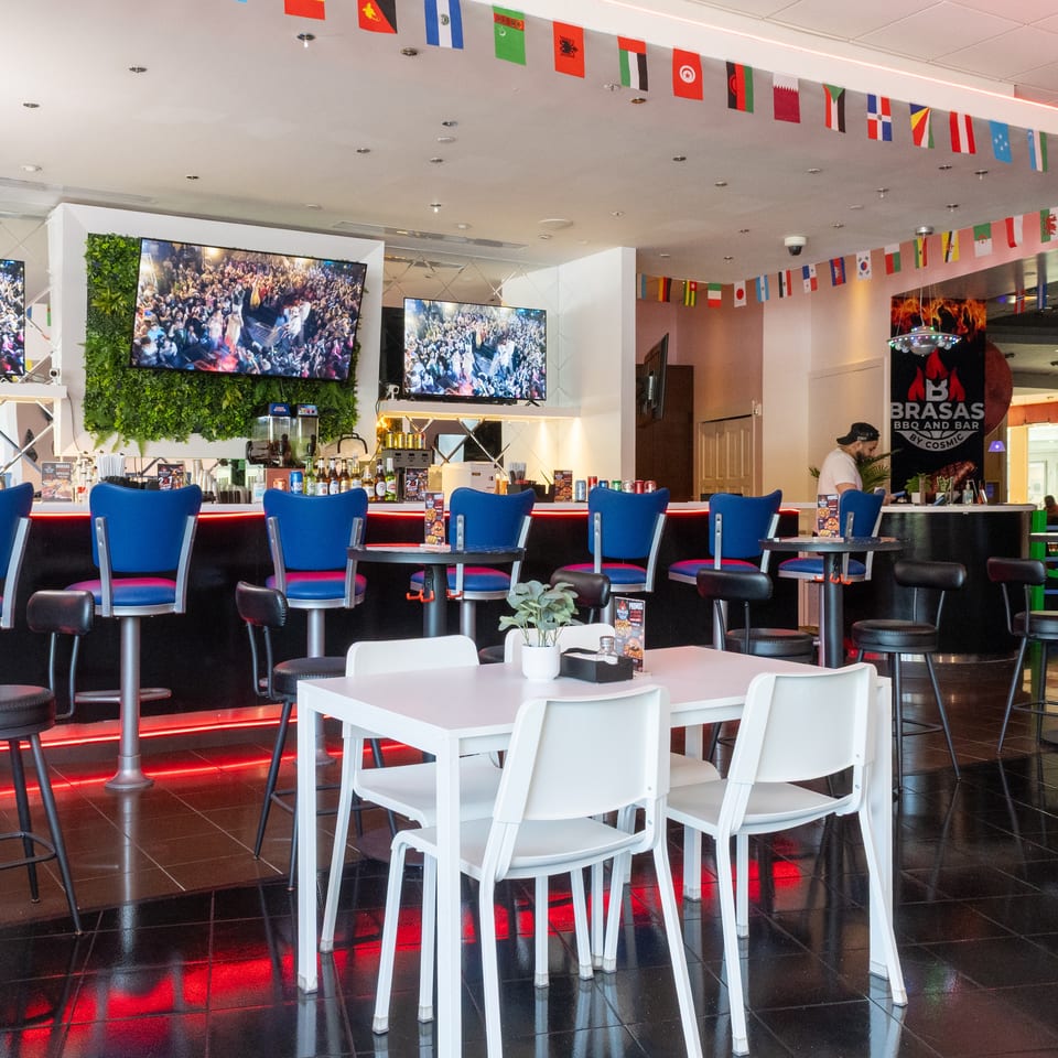 Modern bar area with blue stools, white tables, large TVs, and international flags strung across the ceiling.