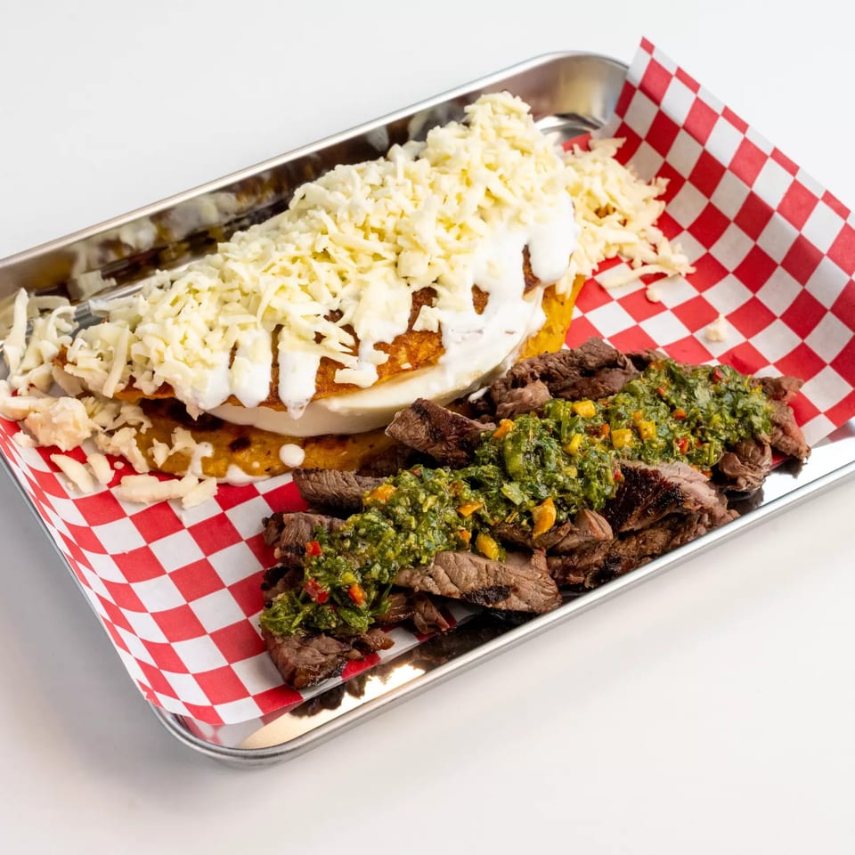 Cheesy baked potato topped with sour cream alongside grilled steak strips with green herb sauce in a checkered-lined metal tray.