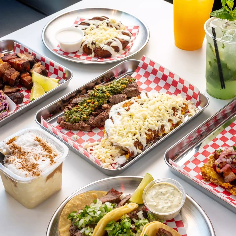 Assorted Latin American dishes, including grilled meats, tacos, cheesy entrées, slaw, sauces, and refreshing drinks, served on a restaurant table.